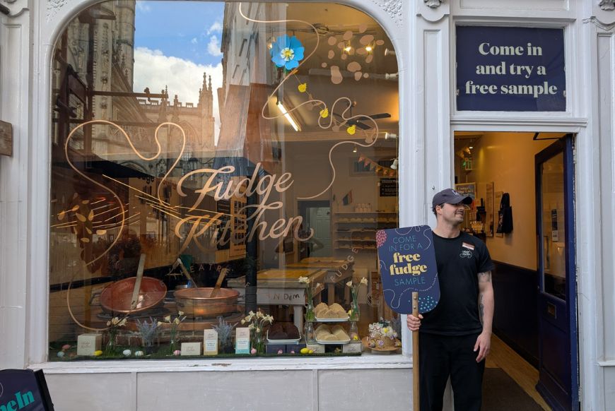 A shop named fudge kitchen, with a worker standing outside holding sign that says free fudge